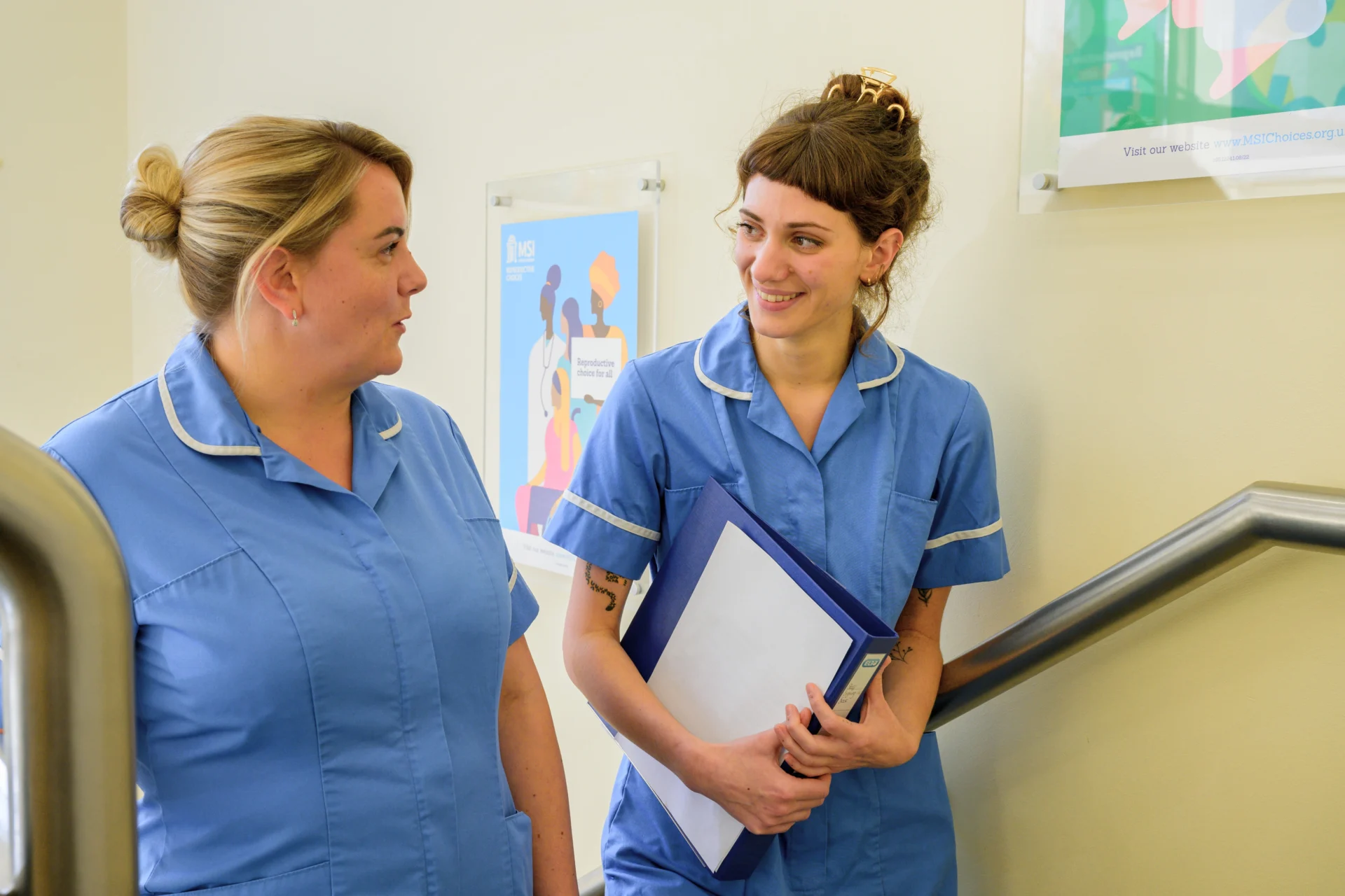 Close-up of two MSI nurses smiling and talking as they walk up the stairs in a clinic.