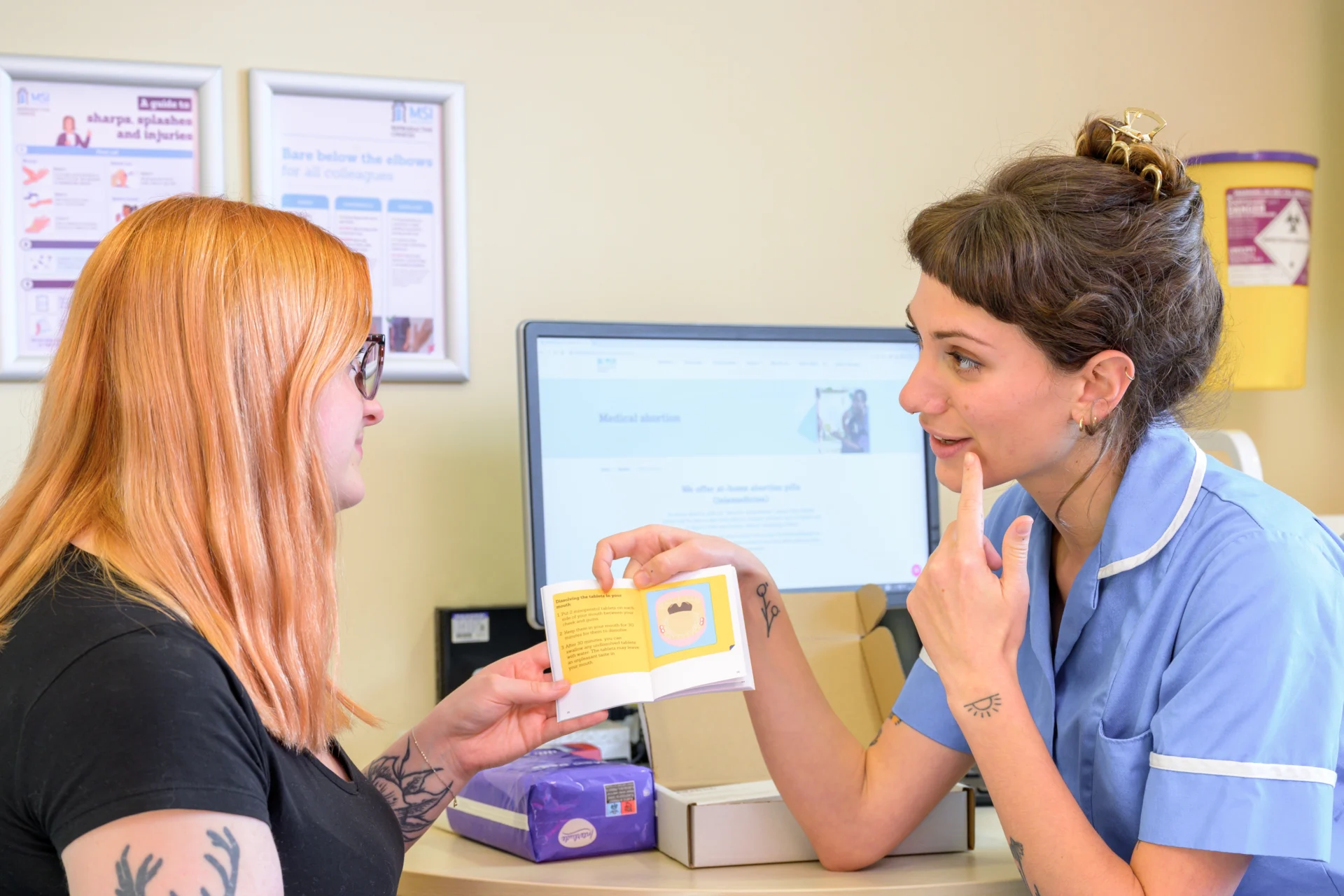An MSI nurse and client sat at a table. The MSI nurse is pointing to their gums whilst holding open the medical abortion booklet on the page explaining how to take the abortion tablets.